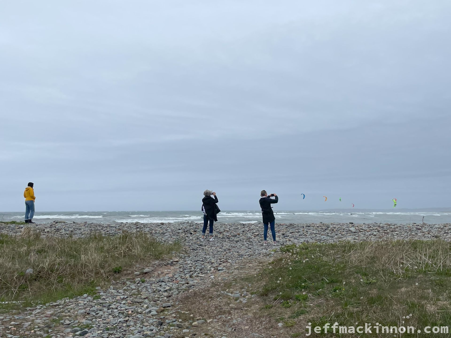 Three people looking out over the water at kite surfers, two with cameras.