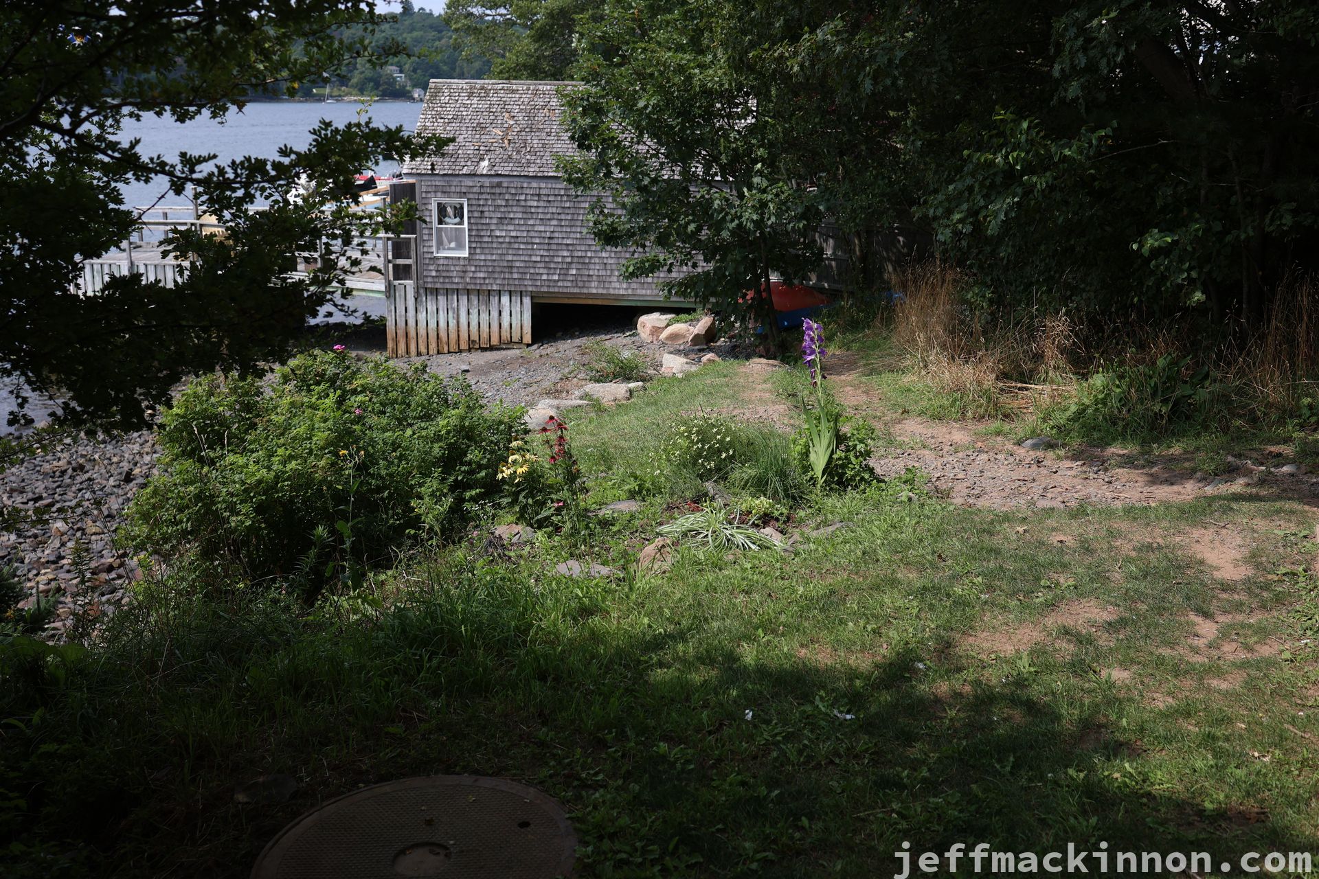 A dirt path going down towards the water with a boat house to the right and a small flower garden to the left.